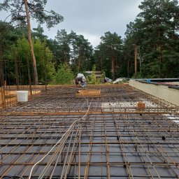 USŁUGI REMONTOWO BUDOWLANE - Zbrojenie płyty fundamentowej domu jednorodzinnego z widocznym pracownikiem w tle, siatka zbrojeniowa ułożona na czarnej folii, widok z góry na las i pochmurne niebo.
