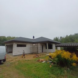 USŁUGI REMONTOWO BUDOWLANE - Budowa parterowego domu z bloczków, z dachem pokrytym papą. Na dachu stoi pracownik, drugi przechodzi obok budynku. Widoczny bus i rusztowanie.