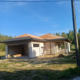 USŁUGI REMONTOWO BUDOWLANE - Budowa parterowego domu z garażem w stanie surowym otwartym, widoczne bloczki, drewniana konstrukcja dachu i rusztowania. Słoneczny dzień, błękitne niebo.