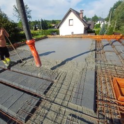 USŁUGI REMONTOWO BUDOWLANE - Wylewanie betonu na strop zbrojony, widoczna pompa do betonu i pracownik wyrównujący powierzchnię. W tle dom jednorodzinny i błękitne niebo.