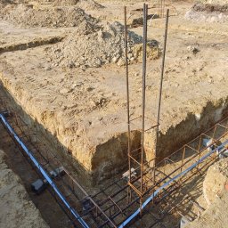 USŁUGI REMONTOWO BUDOWLANE - Zbrojenie fundamentów w wykopie, widoczne stalowe pręty i szalunki. W tle ziemia i fragment ogrodzenia. Ujęcie z góry, oświetlenie naturalne.