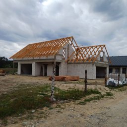 USŁUGI REMONTOWO BUDOWLANE - Surowy stan domu z betonu komórkowego z drewnianą konstrukcją dachu w trakcie budowy, widok z zewnątrz na tle pochmurnego nieba. Częściowo ogrodzony teren.