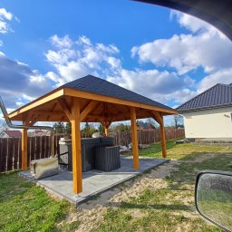 USŁUGI REMONTOWO BUDOWLANE - Drewniana altana z czarnym dachem, osłaniająca szarą balię, na tle błękitnego nieba z chmurami. Widok zza szyby samochodu.