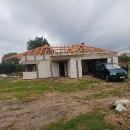 USŁUGI REMONTOWO BUDOWLANE - Niedokończony dom z pustaków z drewnianą konstrukcją dachu w trakcie budowy, na tle pochmurnego nieba. Obok zaparkowany zielony van.