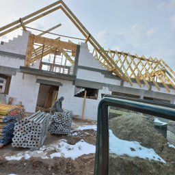 USŁUGI REMONTOWO BUDOWLANE - Budowa domu z białych bloczków: pracownik układa stemple budowlane, widoczna konstrukcja dachu z drewnianych belek, piasek i resztki śniegu. Częściowy widok zza szyby.