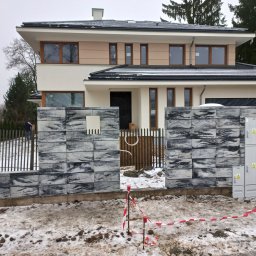 USŁUGI REMONTOWO BUDOWLANE - Nowoczesny dom z szarego kamienia i drewnianym ogrodzeniem w zimowej scenerii. Widoczne skrzynki elektryczne i taśma ostrzegawcza.