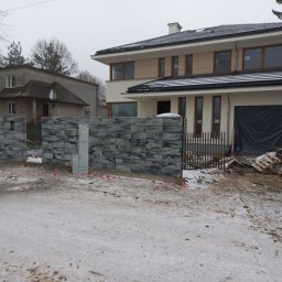 USŁUGI REMONTOWO BUDOWLANE - Nowoczesny, szary ogrodzenie panelowe z widoczną skrzynką elektryczną na tle domu w trakcie budowy, częściowo zasłoniętego czarną folią. Zimowy krajobraz.