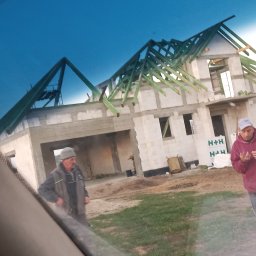 USŁUGI REMONTOWO BUDOWLANE - Budowa domu z bloczków H+H w trakcie realizacji, widoczna konstrukcja dachu i dwóch mężczyzn na placu budowy. Ujęcie zza szyby samochodu.