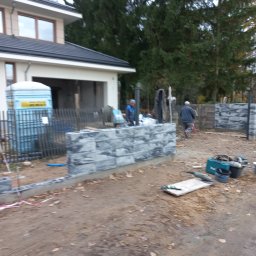 USŁUGI REMONTOWO BUDOWLANE - Wznoszenie ogrodzenia z szarej kostki betonowej na posesji. Widoczne narzędzia, pracownicy w tle i przenośna toaleta. Częściowo ukończona konstrukcja ogrodzenia.