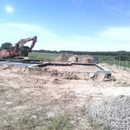 Wykop pod fundamenty domu jednorodzinnego, widoczny ciężki sprzęt budowlany i częściowo zalane fundamenty, teren wyrównany, w tle ogrodzenie i zieleń.