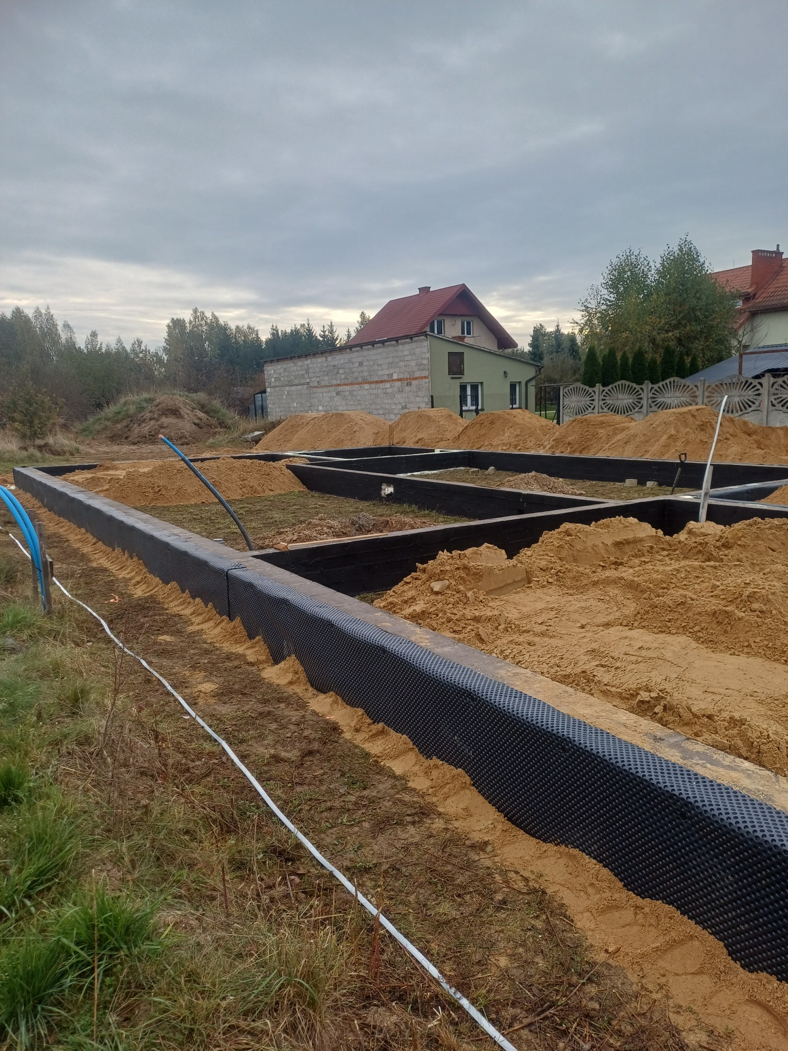 Zabezpieczone fundamenty pod budowę domu, wypełnione piaskiem. Widoczne czarne folie izolacyjne i niebieskie rury. W tle dom w trakcie budowy i pochmurne niebo.