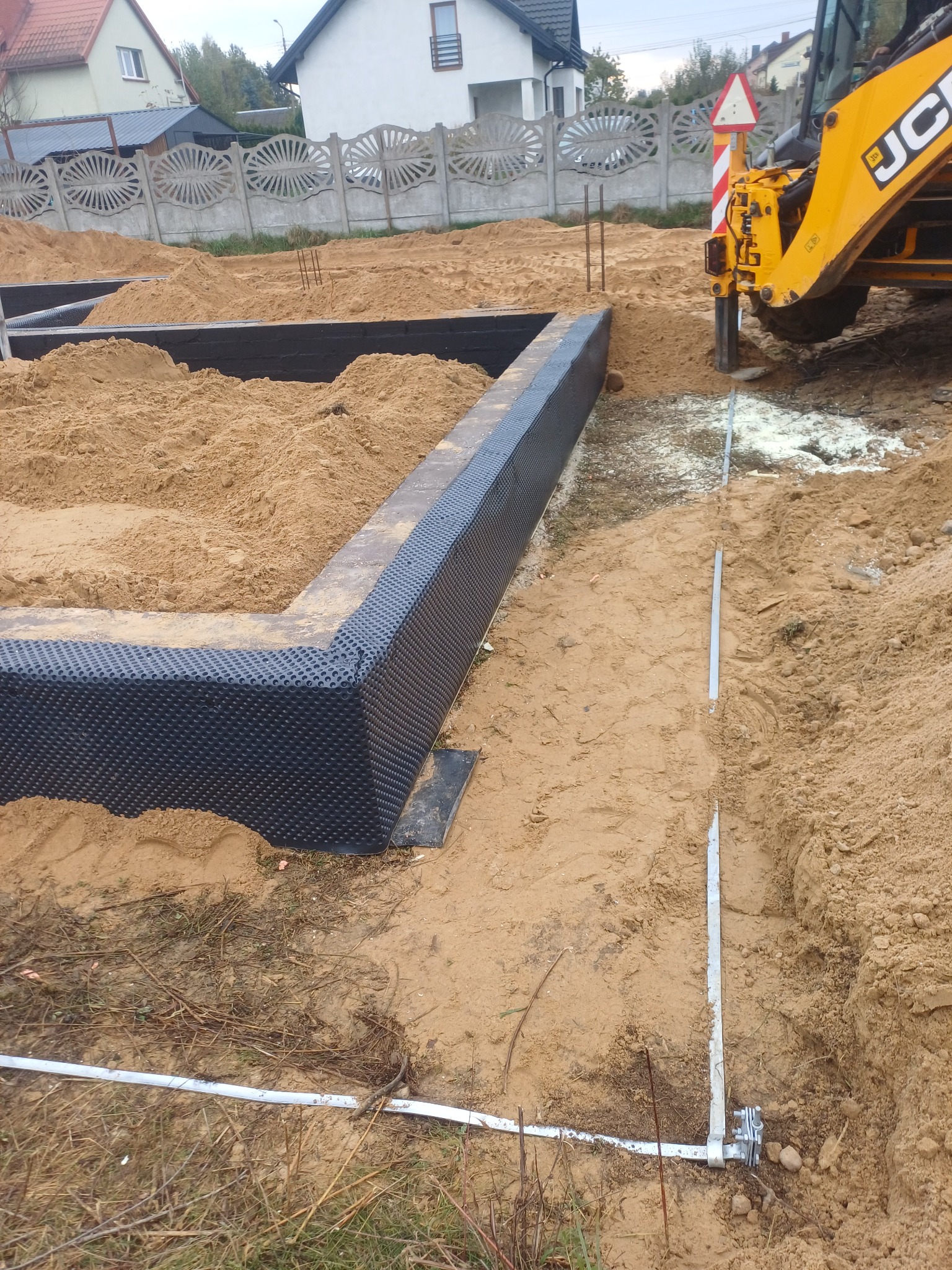 Przygotowany fundament z izolacją pionową, widoczny wykop i piasek. W tle domy i częściowo widoczna koparka JCB.