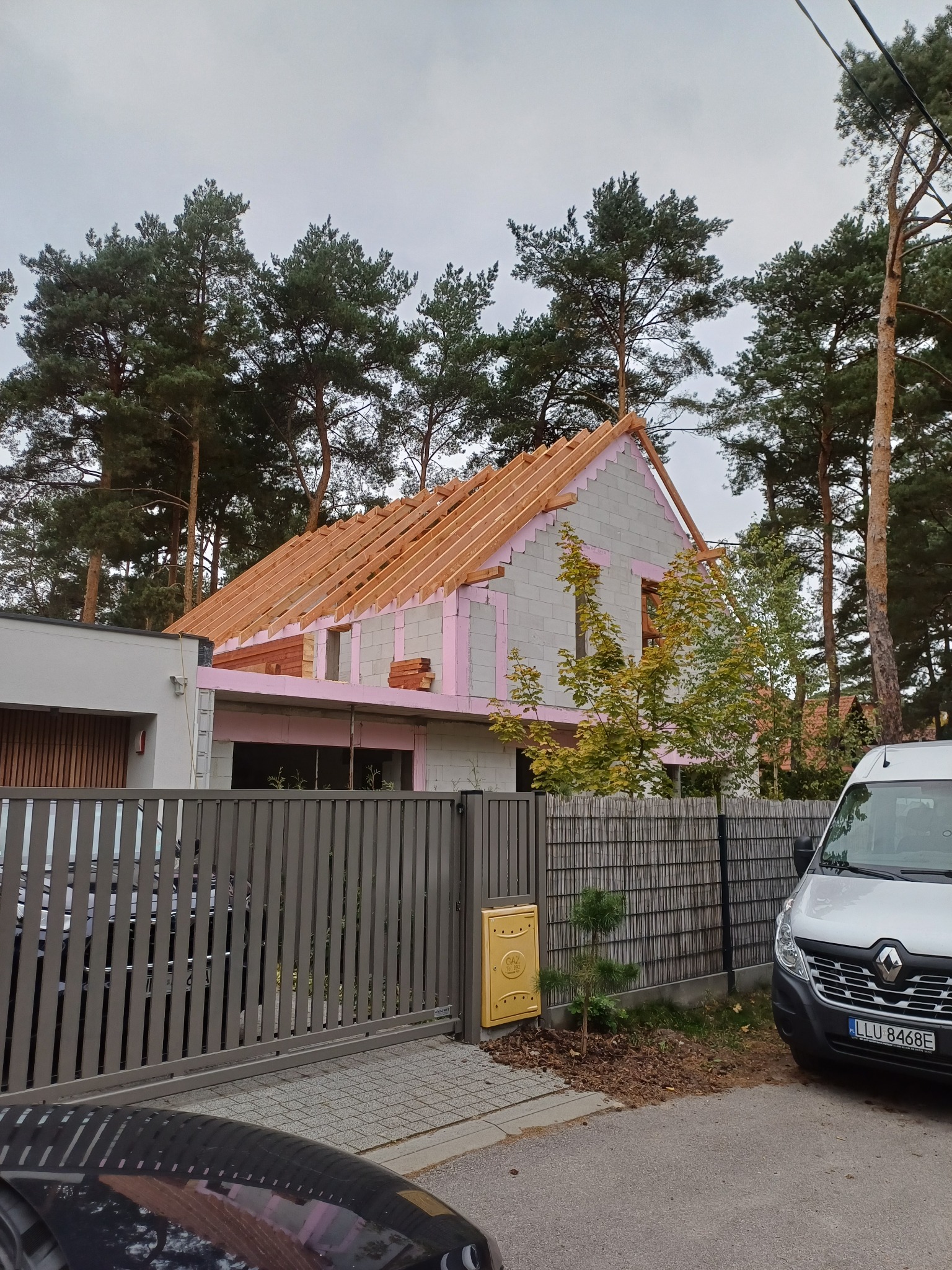 Budowa domu z bloczków, widoczne drewniane elementy konstrukcji dachu i różowe ocieplenie. Na pierwszym planie ogrodzenie i samochód dostawczy.