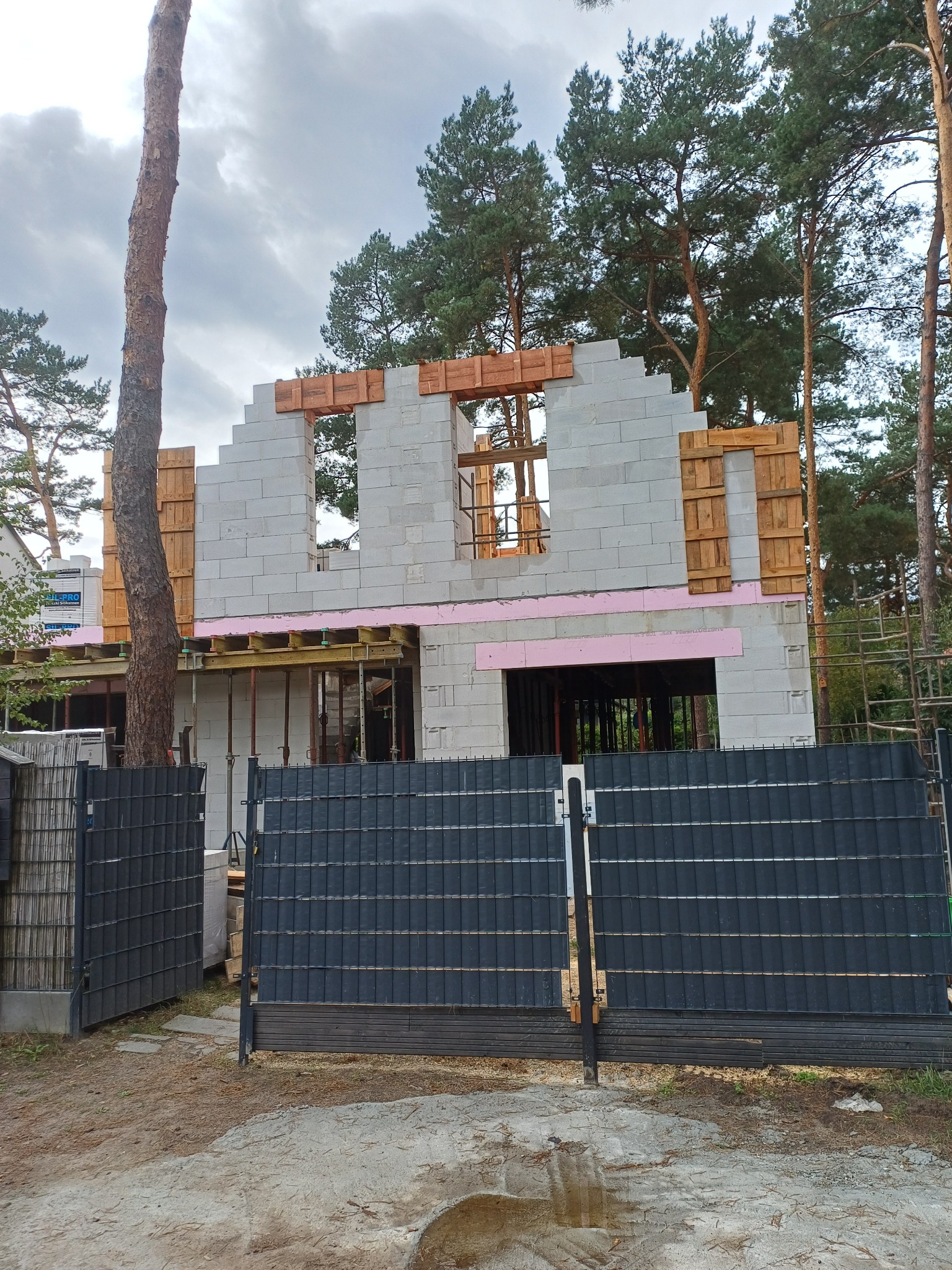 Budowa domu z betonu komórkowego w lesie, widoczne drewniane szalunki okien i różowa izolacja. W tle wysokie drzewa i fragment ogrodzenia.