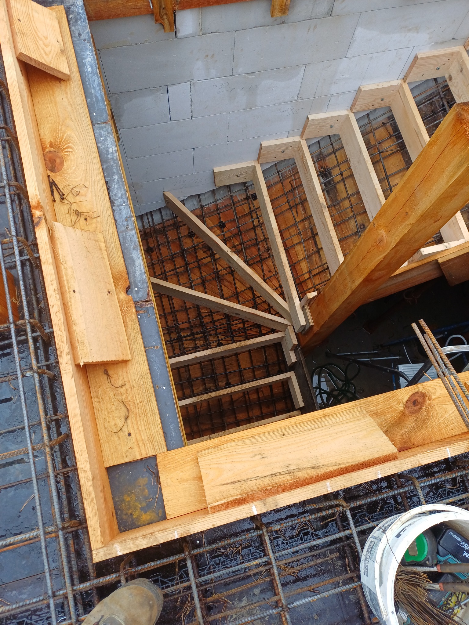 Zbrojenie schodów z drewnianym szalunkiem i siatką zbrojeniową, widok z góry. Elementy konstrukcyjne w trakcie budowy, z widocznym fragmentem buta pracownika.