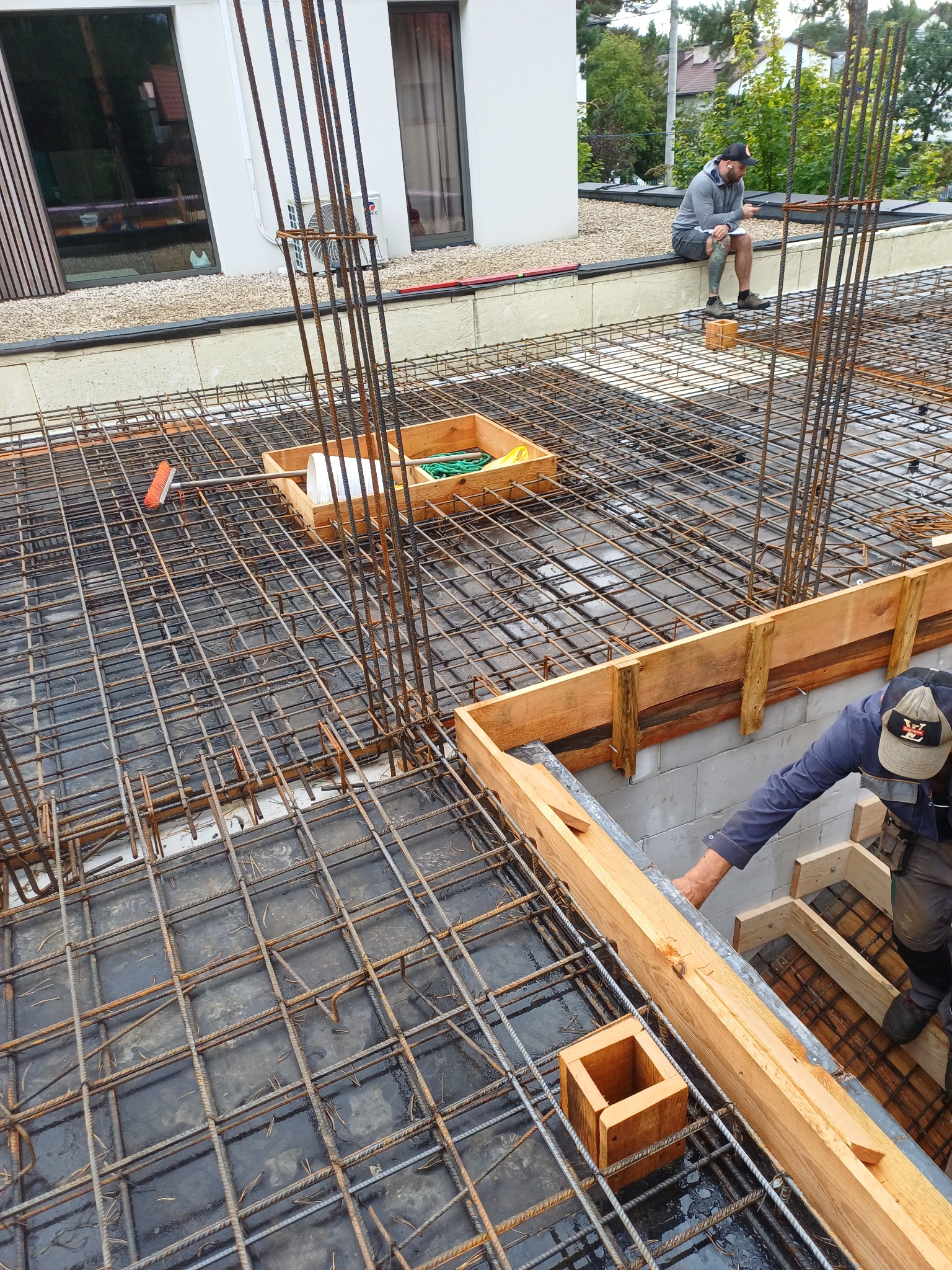 Zbrojenie fundamentów z drewnianym szalunkiem, widoczne pręty zbrojeniowe i pracownik przy szalunku. W tle budynek z oknami i osoba siedząca na murku.