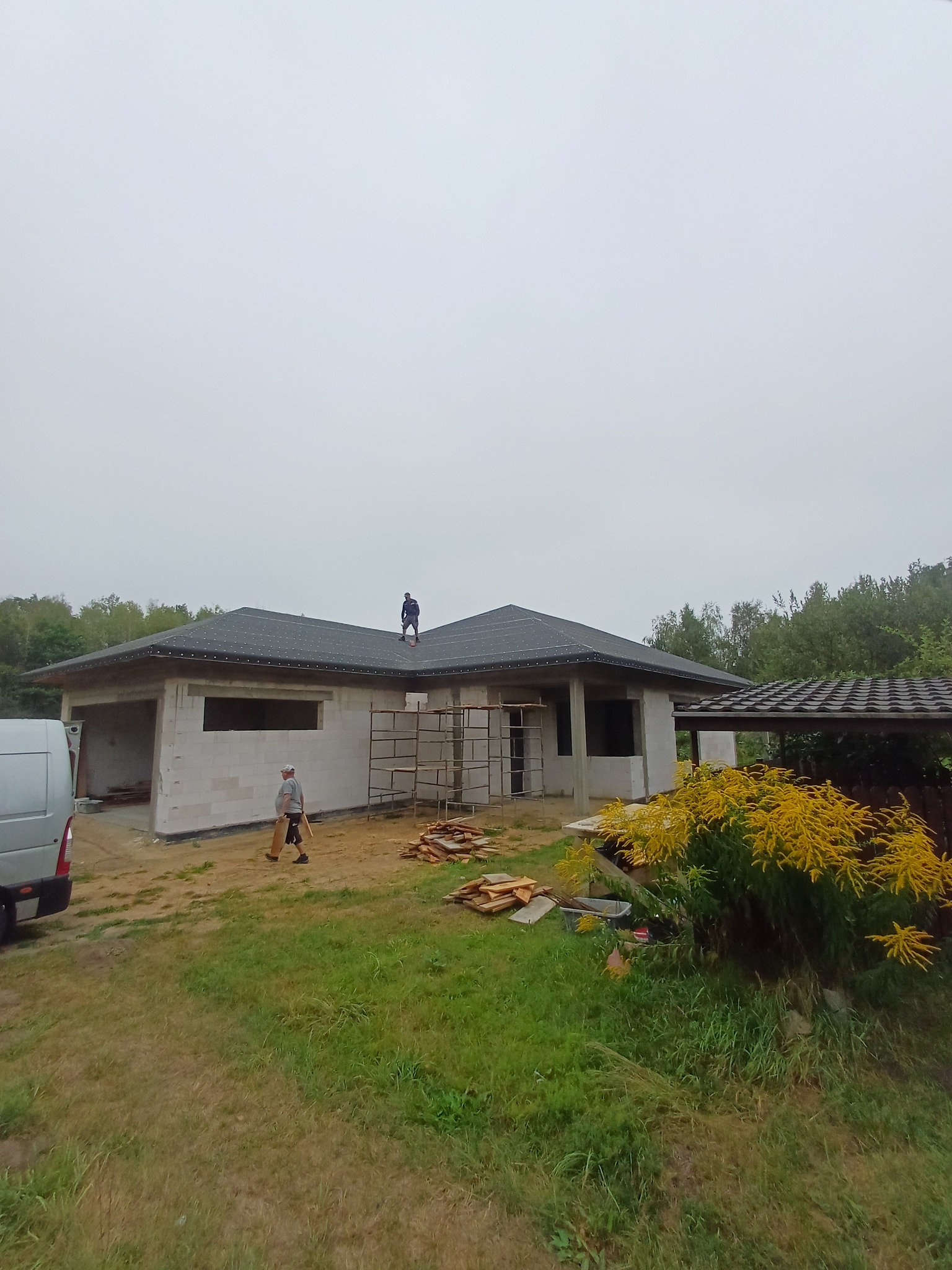 Budowa parterowego domu z bloczków, z dachem pokrytym papą. Na dachu stoi pracownik, drugi przechodzi obok budynku. Widoczny bus i rusztowanie.