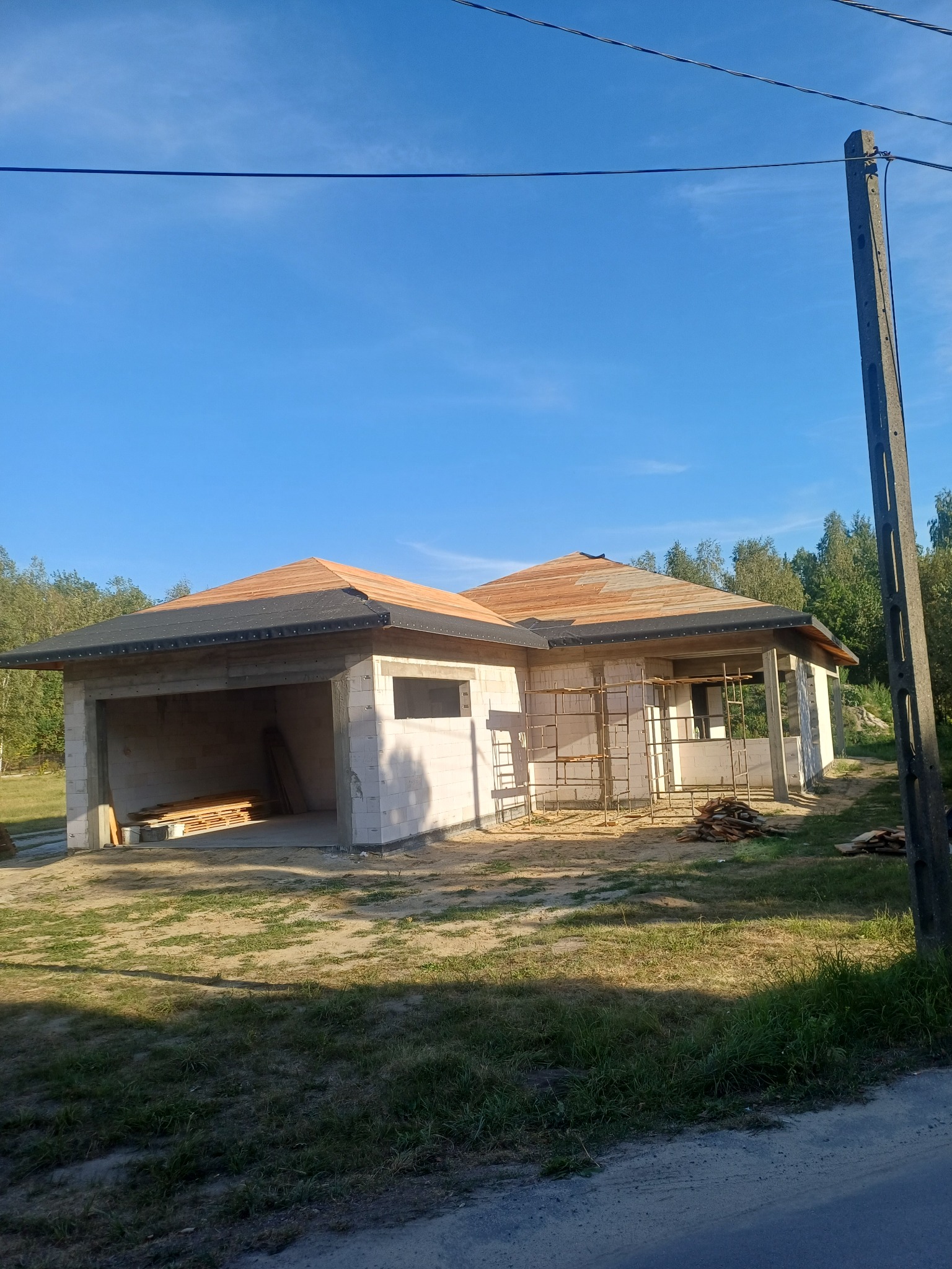 Budowa parterowego domu z garażem w stanie surowym otwartym, widoczne bloczki, drewniana konstrukcja dachu i rusztowania. Słoneczny dzień, błękitne niebo.