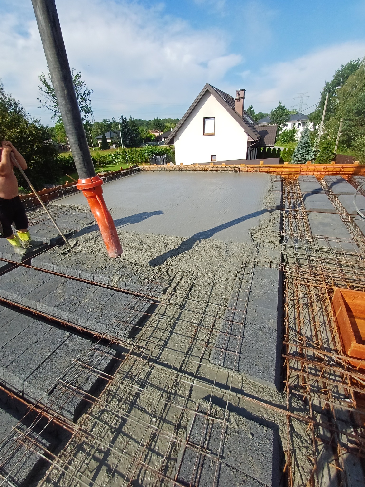 Wylewanie betonu na strop zbrojony, widoczna pompa do betonu i pracownik wyrównujący powierzchnię. W tle dom jednorodzinny i błękitne niebo.