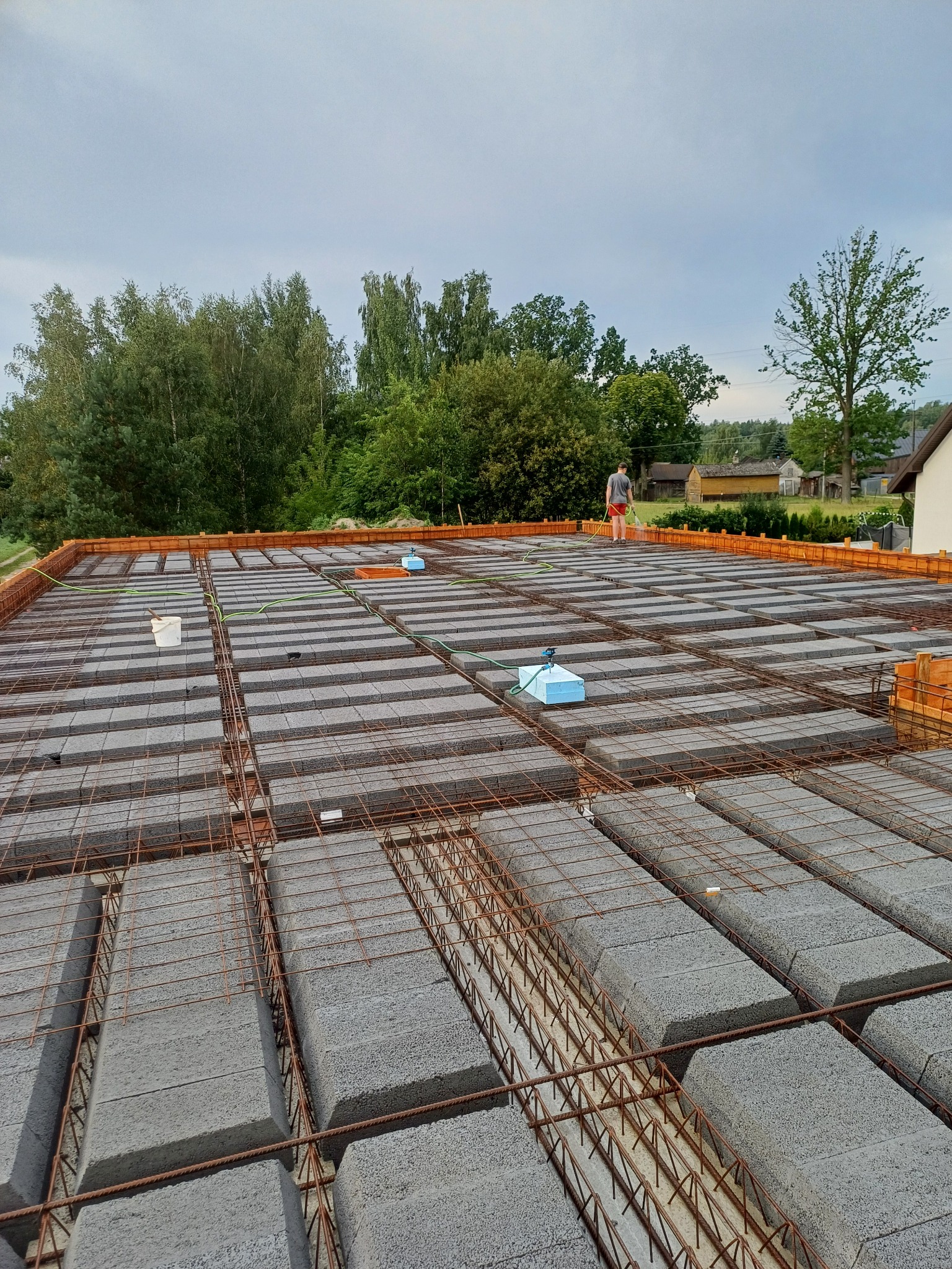 Zbrojona konstrukcja stropu Teriva z betonowych pustaków, widok z góry, na tle zieleni i pochmurnego nieba. Osoba podlewa konstrukcję.