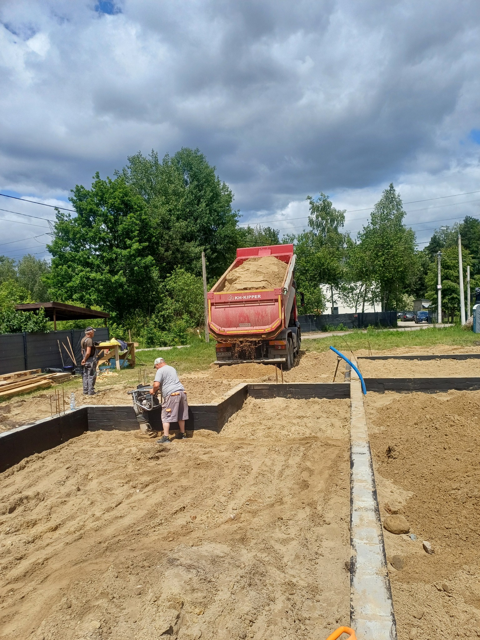 Wyrównywanie piasku w szalunku fundamentów pod budowę domu. Czerwona wywrotka z piaskiem w tle. Pracownik zagęszcza piasek ubijarką.