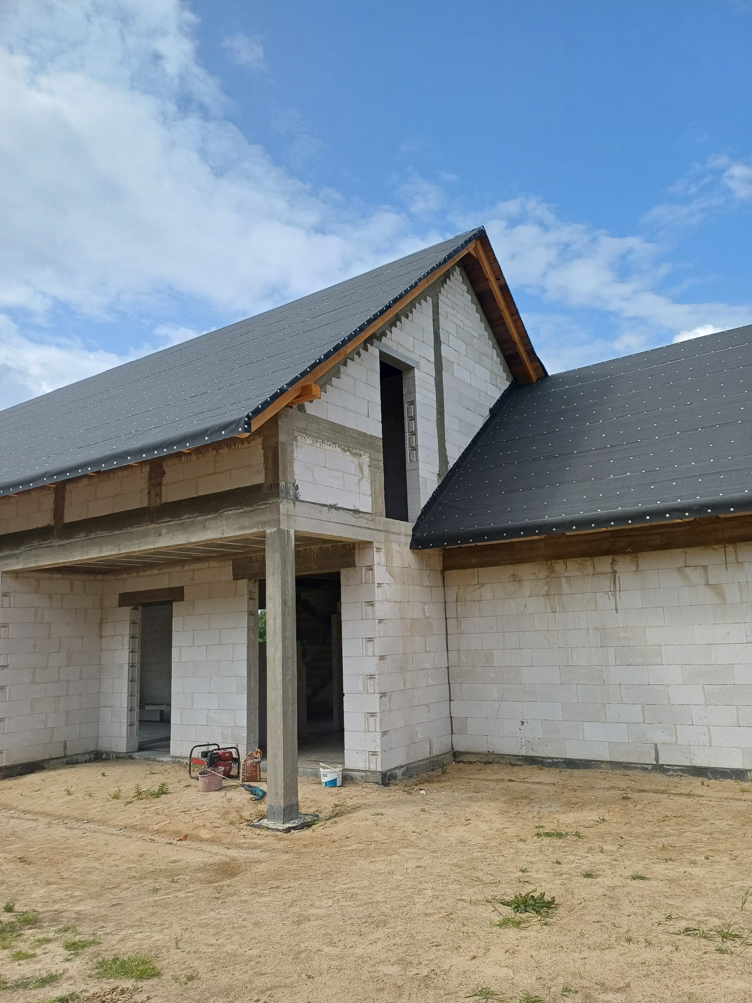 Surowy stan domu z białego bloczka, zadaszony czarną papą, widoczne betonowe elementy konstrukcyjne, teren wokół w stanie budowy, błękitne niebo w tle.