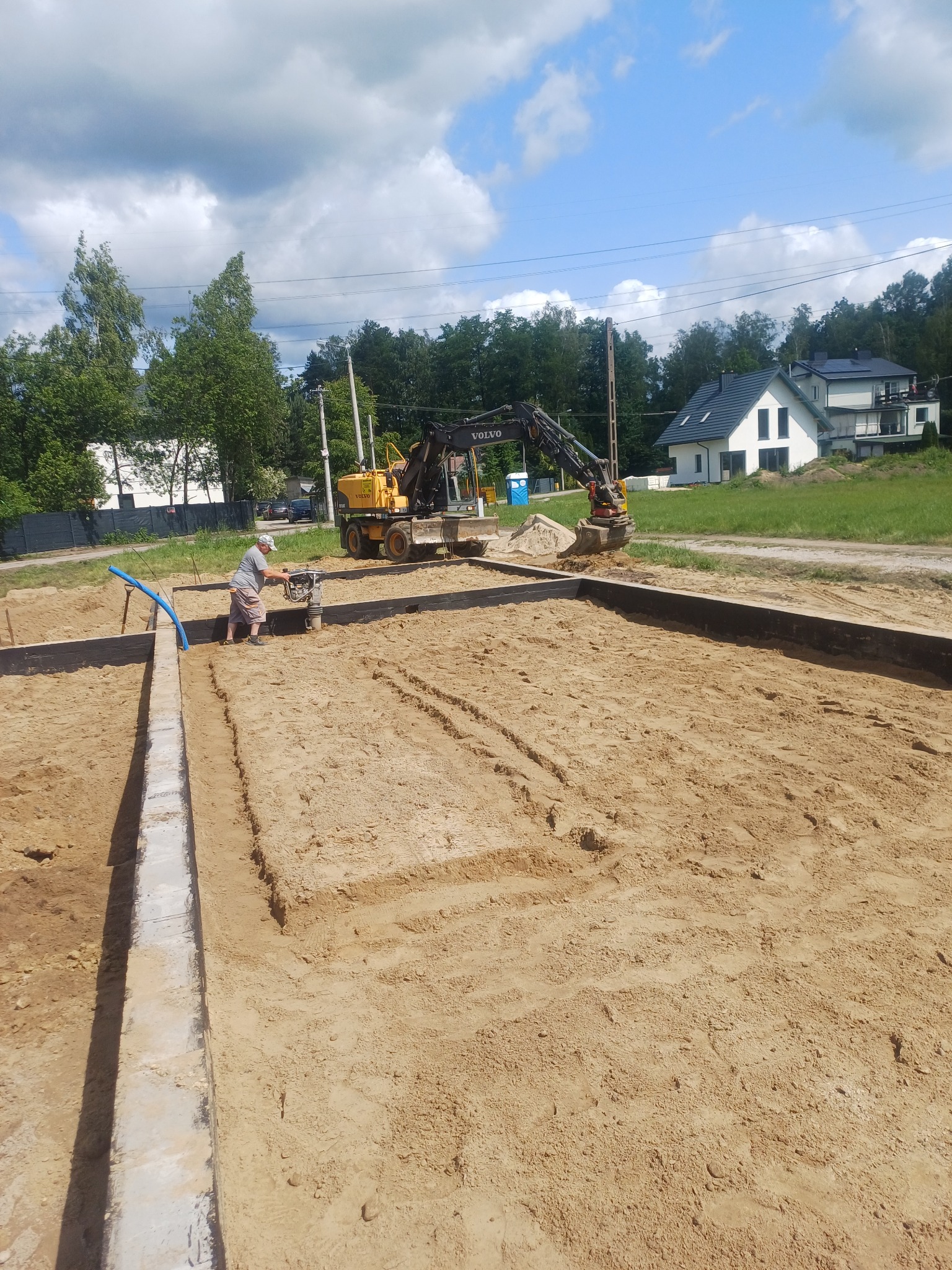 Przygotowanie terenu pod fundamenty: pracownik zagęszcza piasek ubijarką, w tle wykop i koparka Volvo, widoczne szalunki. Prace ziemne w słoneczny dzień.
