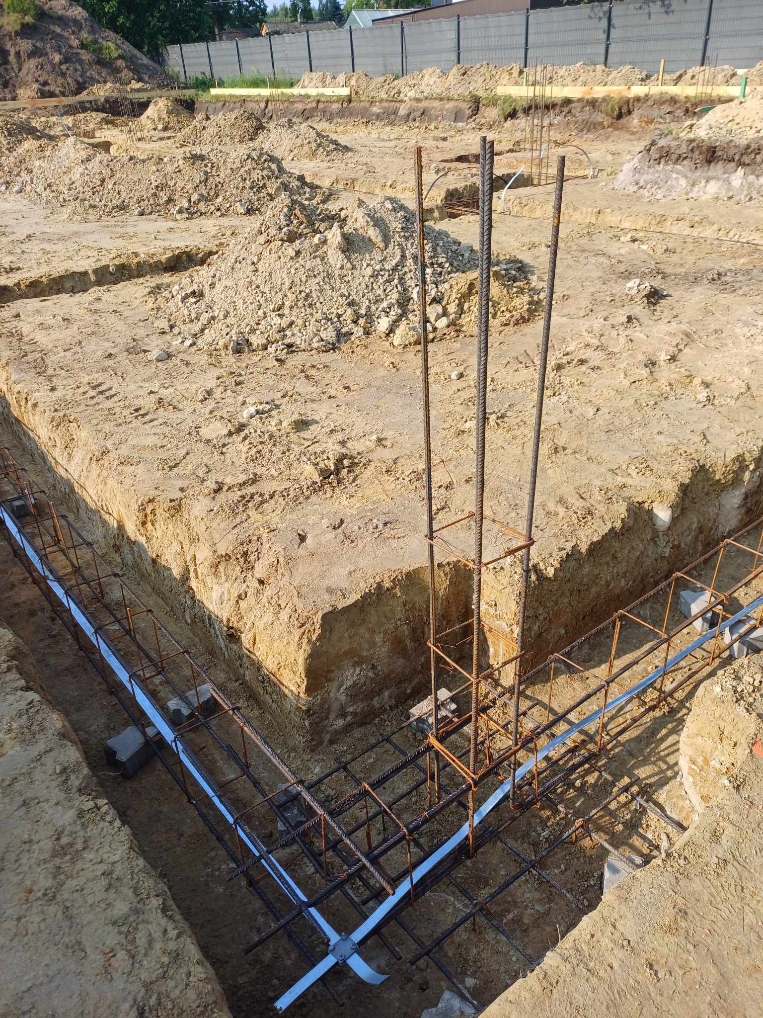 Zbrojenie fundamentów w wykopie, widoczne stalowe pręty i szalunki. W tle ziemia i fragment ogrodzenia. Ujęcie z góry, oświetlenie naturalne.