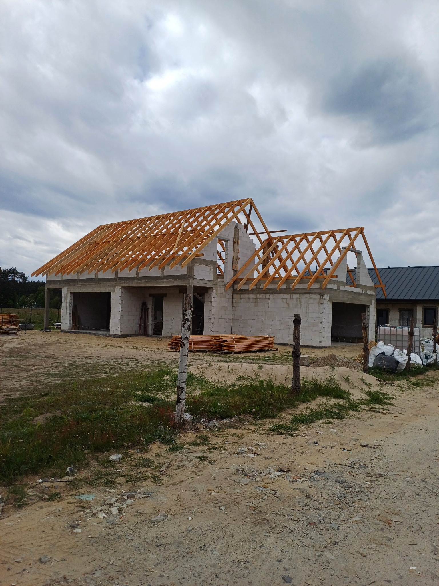 Surowy stan domu z betonu komórkowego z drewnianą konstrukcją dachu w trakcie budowy, widok z zewnątrz na tle pochmurnego nieba. Częściowo ogrodzony teren.