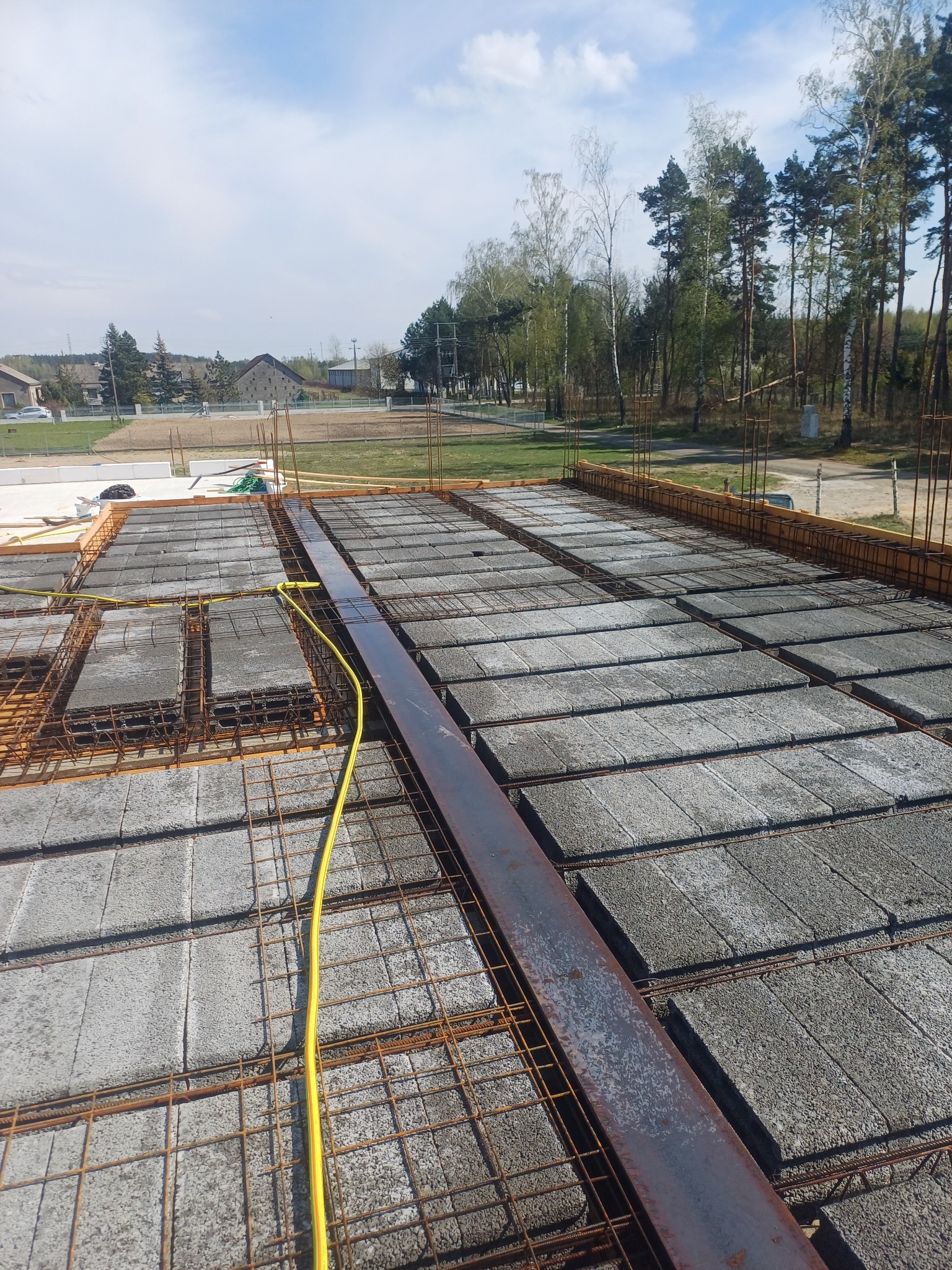 Zbrojenie stropu Teriva z belką stalową i przewodami elektrycznymi, widok z góry na tle zabudowy i drzew. Elementy szalunku widoczne w tle.