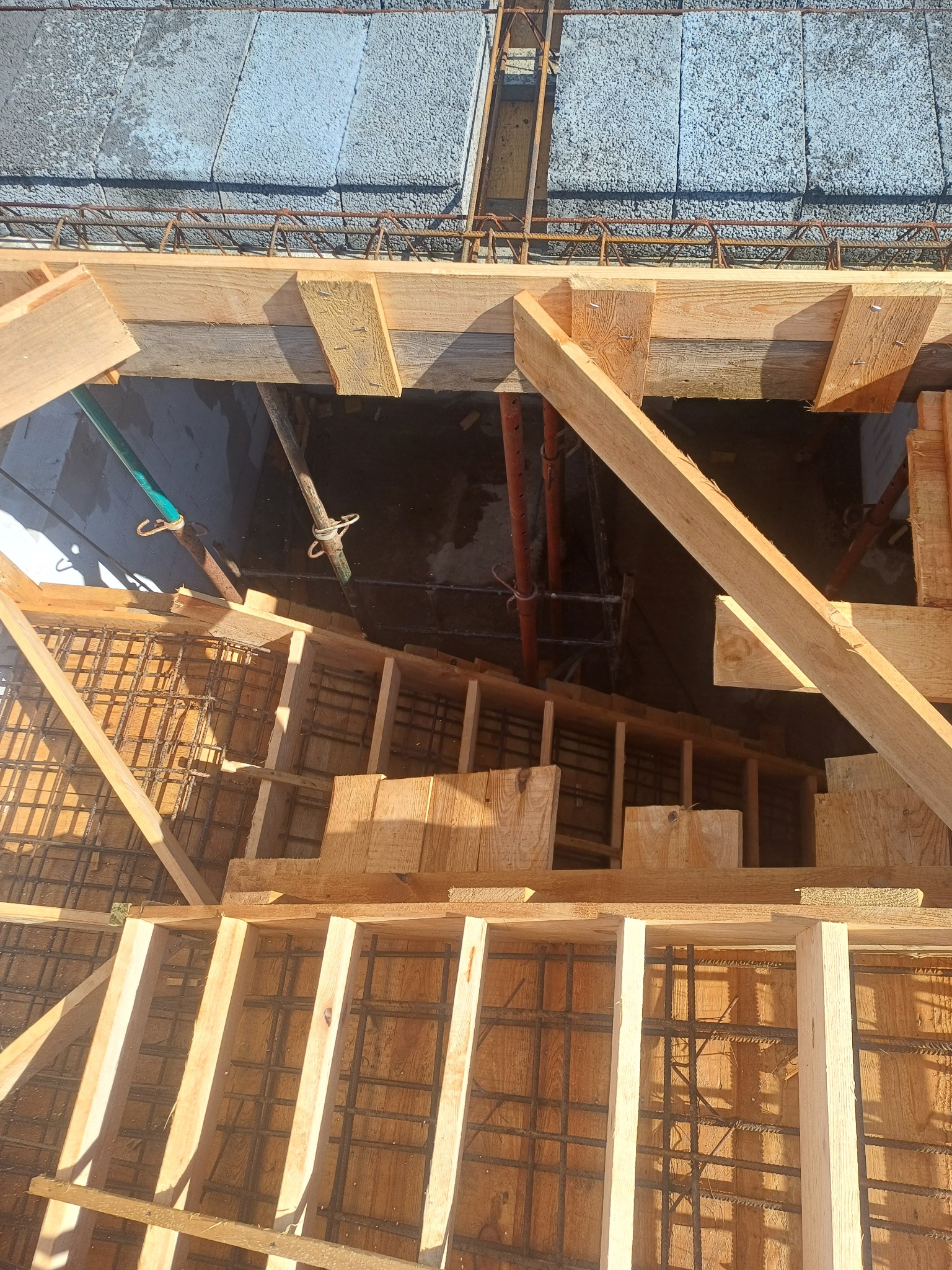 Zbrojenie fundamentów w drewnianym szalunku, widok z góry. Stalowe pręty tworzą siatkę, a drewniane deski formują ściany. Widoczne elementy konstrukcyjne i betonowe bloczki.