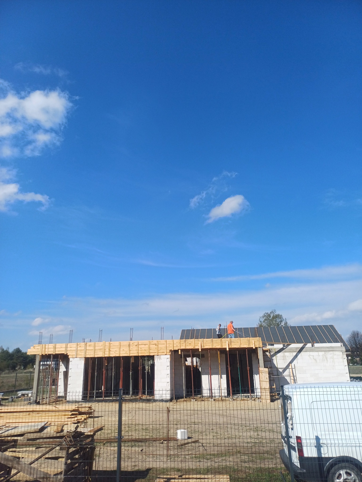 Budowa domu z betonu komórkowego w Tuchowicach, widok zza ogrodzenia. Widoczne zbrojenia, drewniane szalunki i pracownicy na dachu. Błękitne niebo z chmurami w tle.