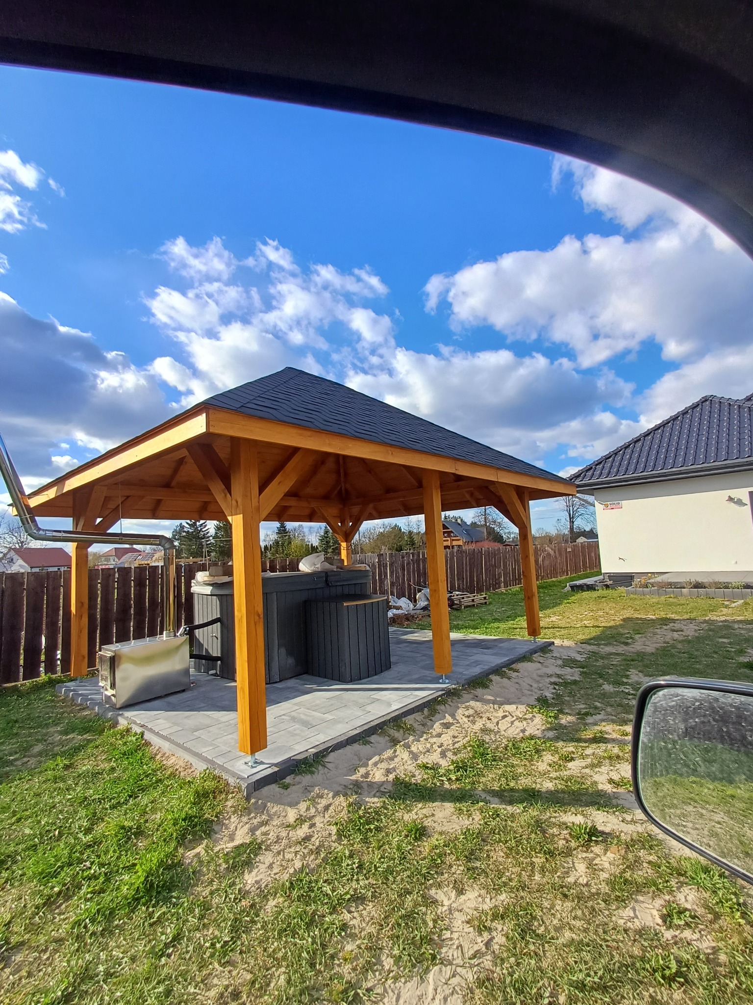 Drewniana altana z czarnym dachem, osłaniająca szarą balię, na tle błękitnego nieba z chmurami. Widok zza szyby samochodu.