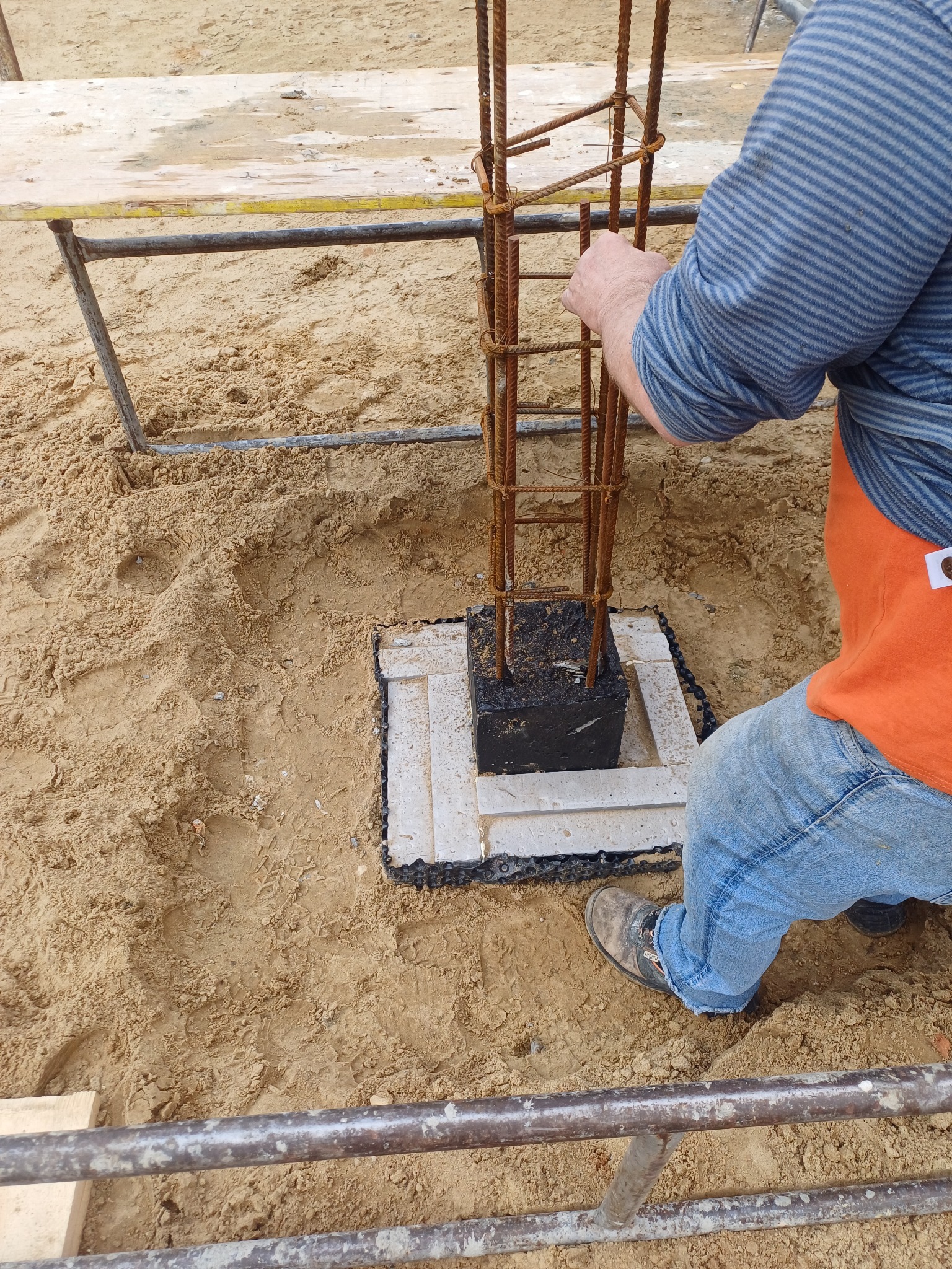 Zbrojenie fundamentu kolumnowego z ułożoną hydroizolacją i drenażem, przygotowane do zalania betonem. Widoczna ręka zbrojarza przy pracy.