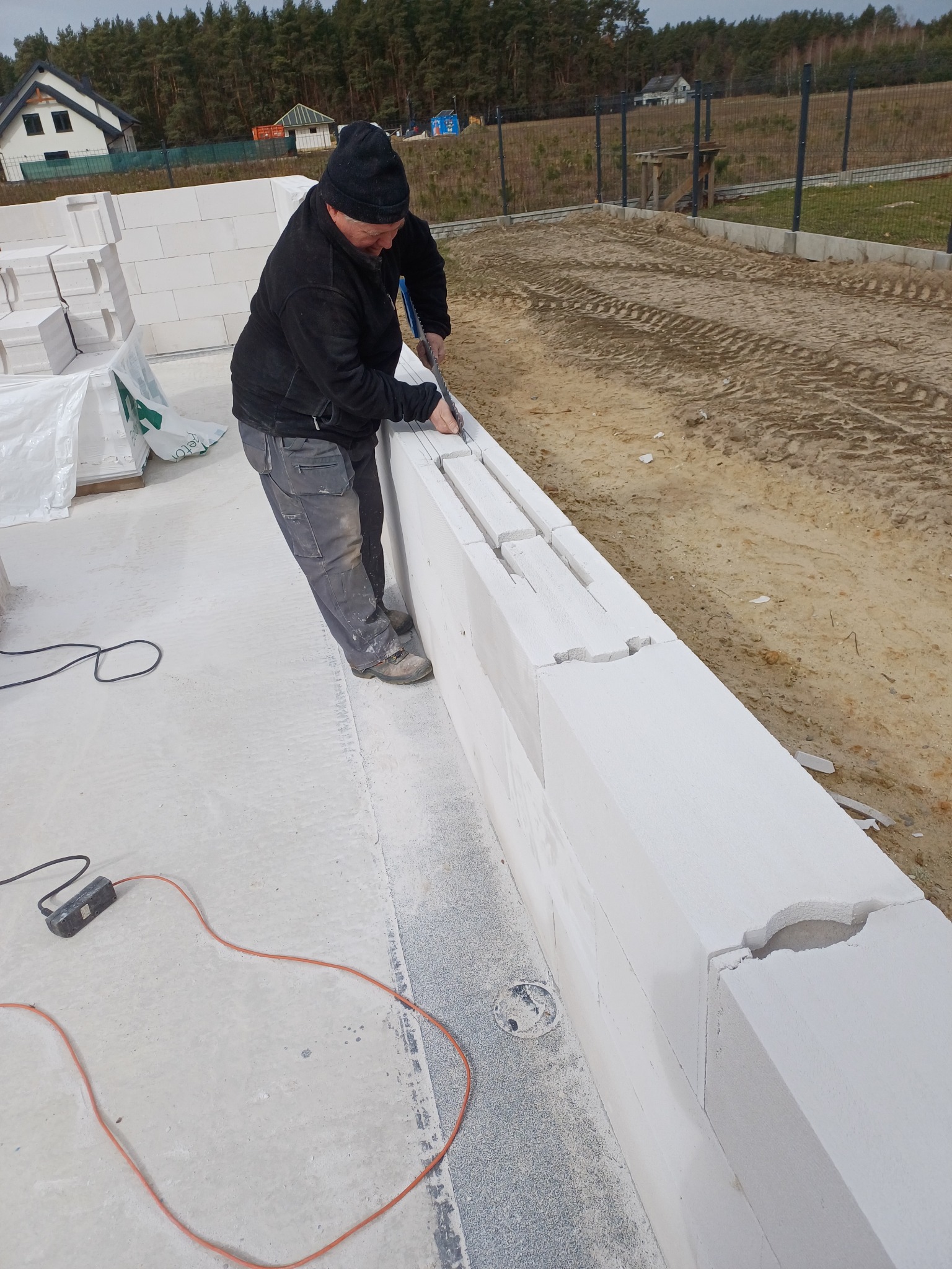 Pracownik docina bloczek z betonu komórkowego piłą ręczną podczas budowy ściany fundamentowej. W tle widoczny teren budowy i dom.