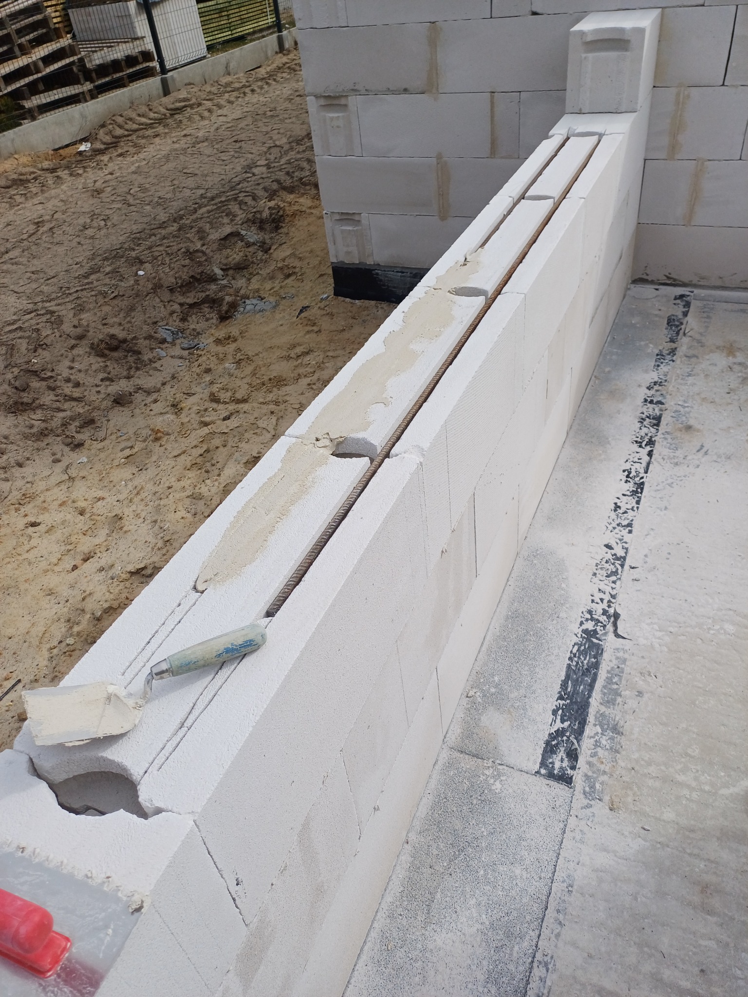 Wznoszenie ściany z betonu komórkowego z widocznym zbrojeniem i kielnią. Ściana w trakcie budowy, z elementami mocującymi i zaprawą.