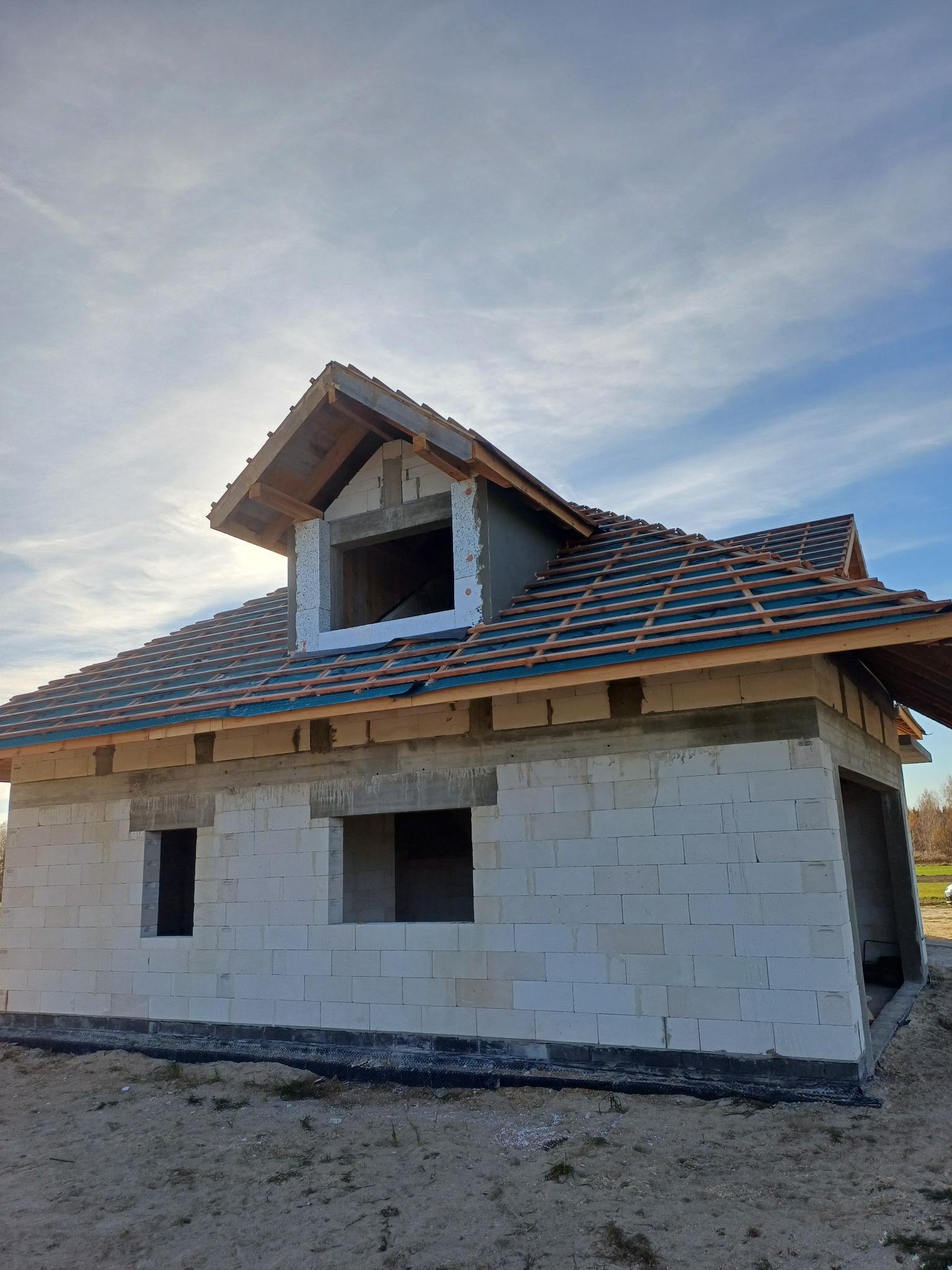 Surowy stan domu z białego pustaka, z częściowo ułożonym dachem z dachówki ceramicznej. Widoczne okna bez ram i lukarna w stanie surowym.