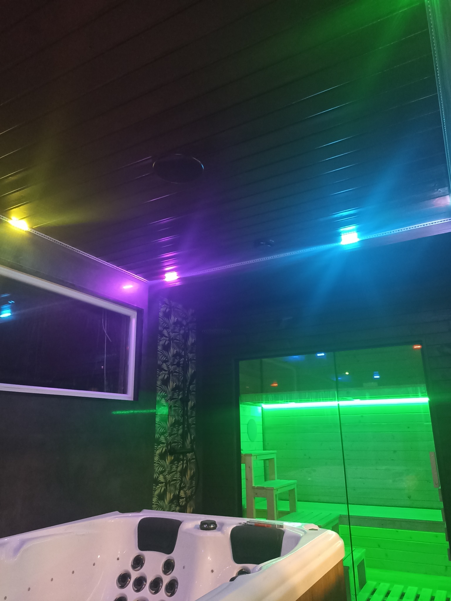 Wnętrze pokoju relaksacyjnego z jacuzzi i sauną, oświetlone kolorowymi diodami LED: żółtymi, fioletowymi i zielonymi. Ściany wykończone ciemnym drewnem, na jednej z nich wzorzysta zasłona. Sufit...