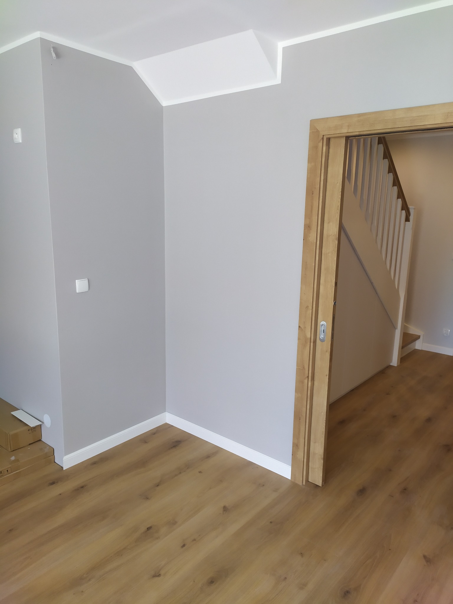 Jasnoszare ściany w nowym mieszkaniu, drewniana podłoga i ościeżnica drzwiowa z widokiem na schody, kartony w rogu