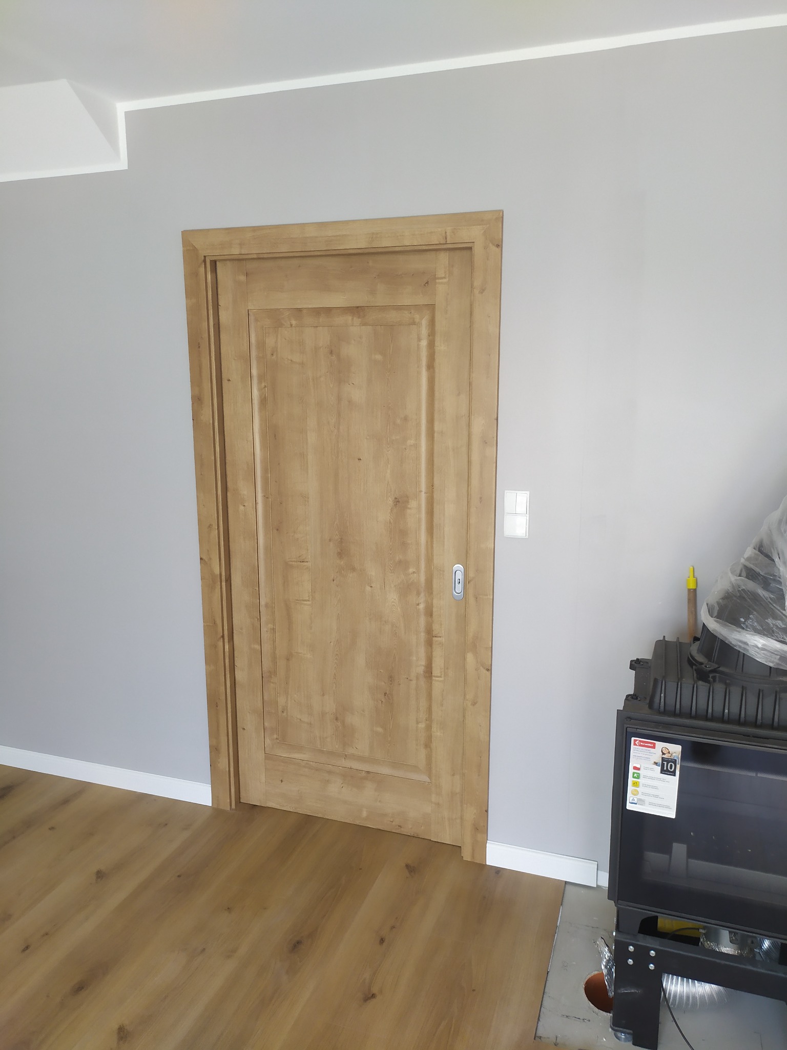 Jasne, drewniane drzwi wewnętrzne z frezowanymi panelami, zamontowane w szarej ścianie z widocznym włącznikiem światła, na podłodze panele imitujące drewno, obok stoi czarny kominek.