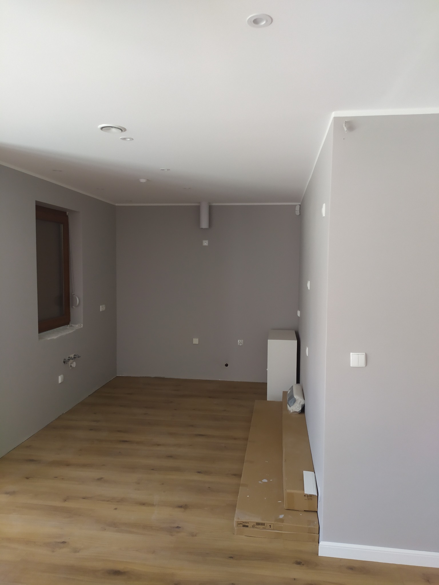 Wnętrze pokoju po remoncie z nową drewnianą podłogą, szarymi ścianami, białym sufitem z oświetleniem punktowym i brązowym oknem. Pod ścianą leżą kartony z panelami.