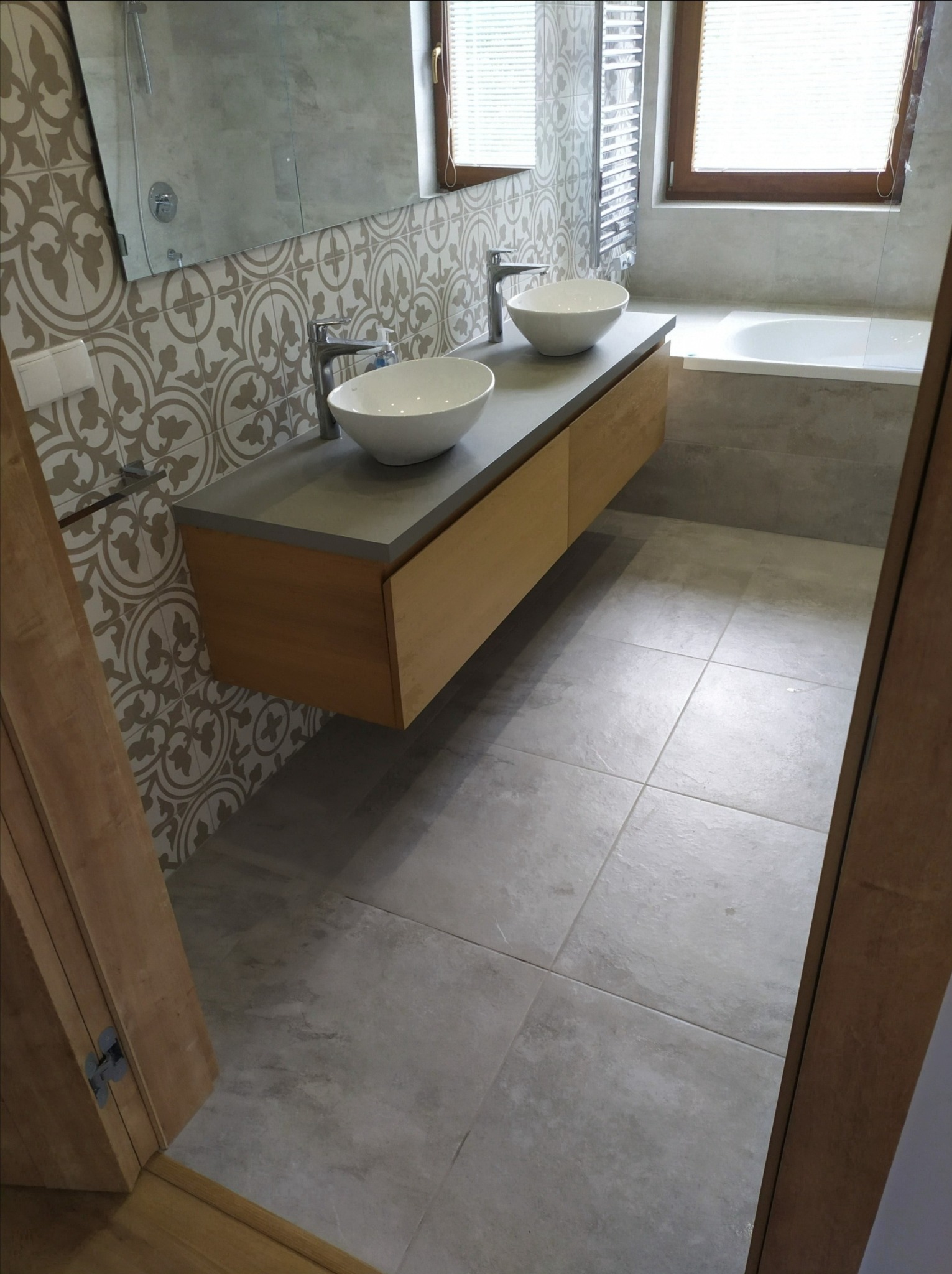 Nowoczesna łazienka z dwoma umywalkami nablatowymi, drewnianą szafką pod umywalką z szarym blatem, wzorzystymi płytkami na ścianie i szarą podłogą z płytek, widok z perspektywy wejścia.