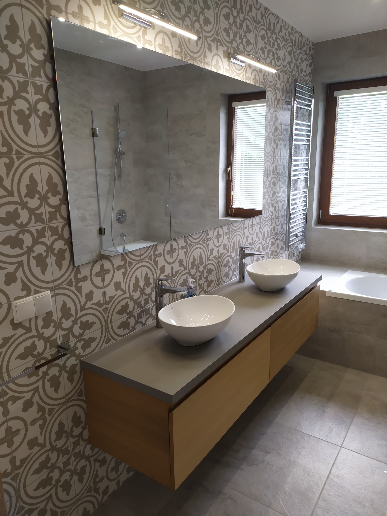 Nowoczesna łazienka z dwoma umywalkami nablatowymi, wzorzystymi płytkami na ścianach i dużym lustrem z oświetleniem LED, widok na grzejnik drabinkowy i okno z żaluzjami.