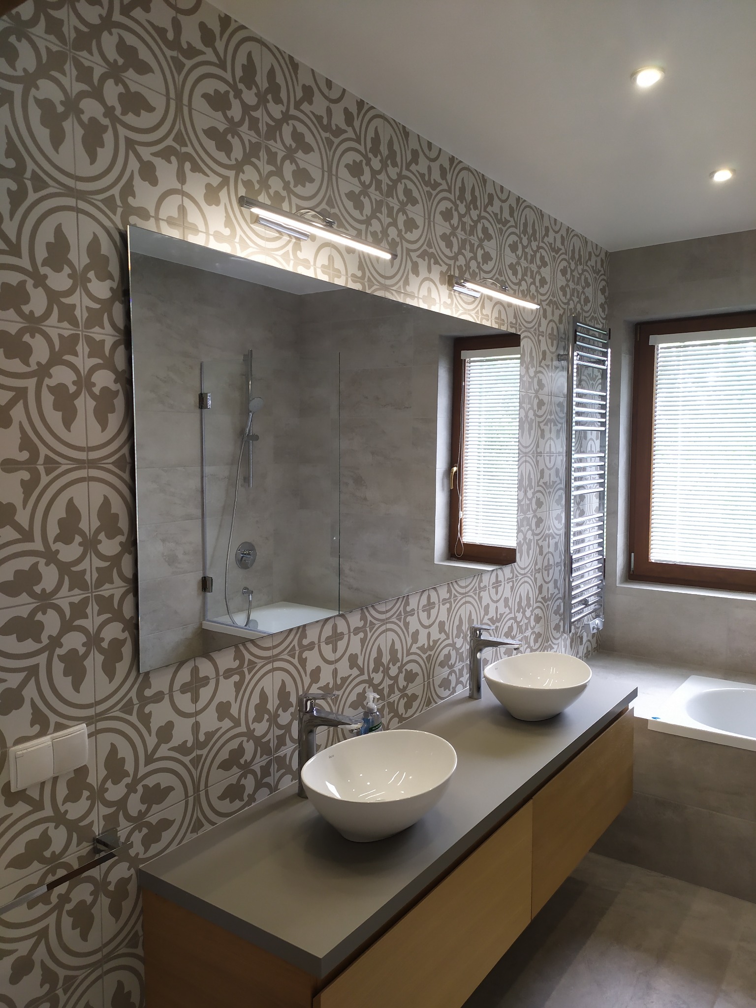 Nowoczesna łazienka z dwoma umywalkami nablatowymi, wzorzystymi płytkami na ścianie i dużym lustrem odbijającym kabinę prysznicową.