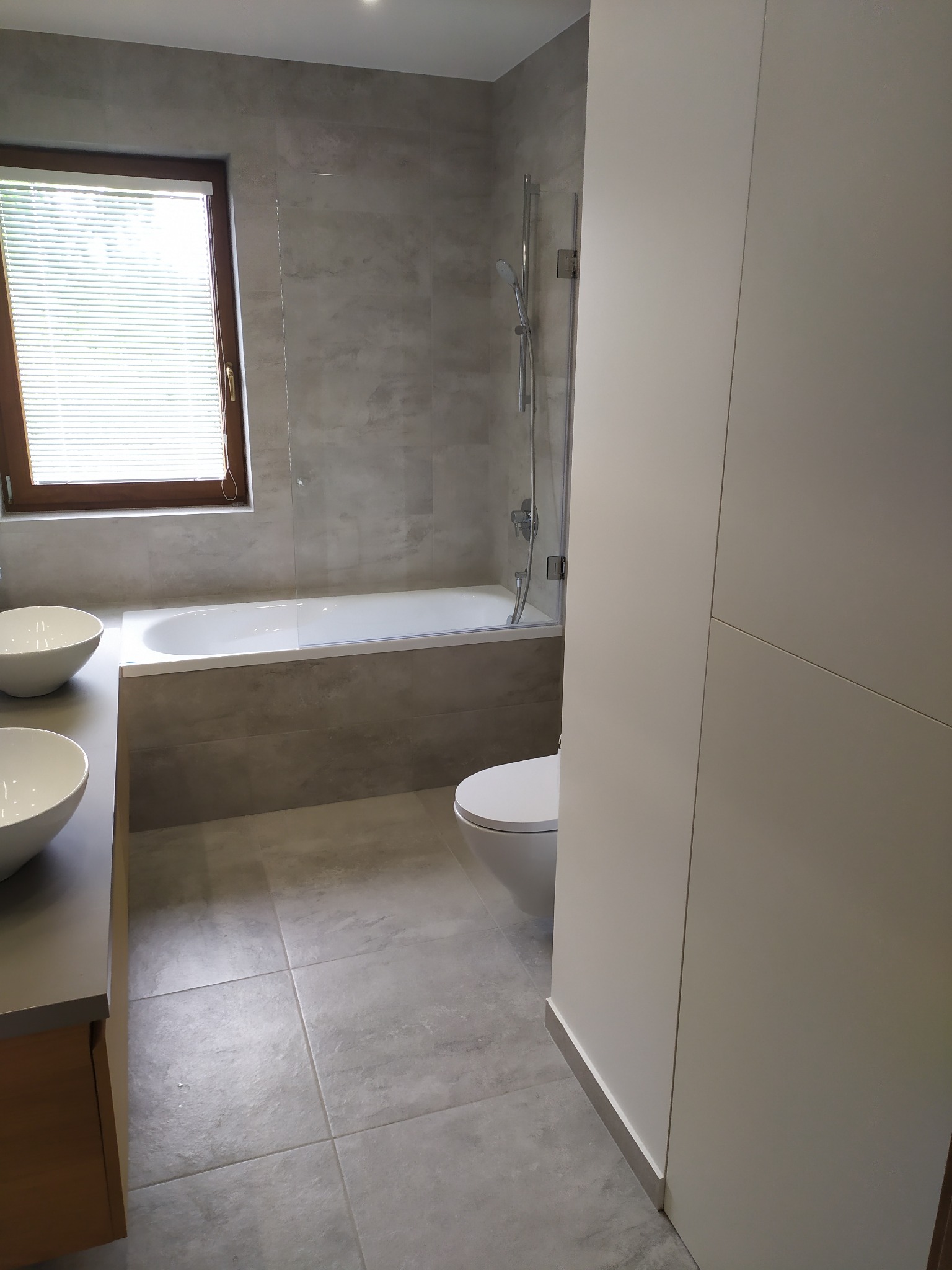 Nowoczesna łazienka z szarymi płytkami, białą wanną i umywalkami nablatowymi w kształcie misy. Widoczne okno z żaluzjami i zabudowa meblowa w kolorze białym.