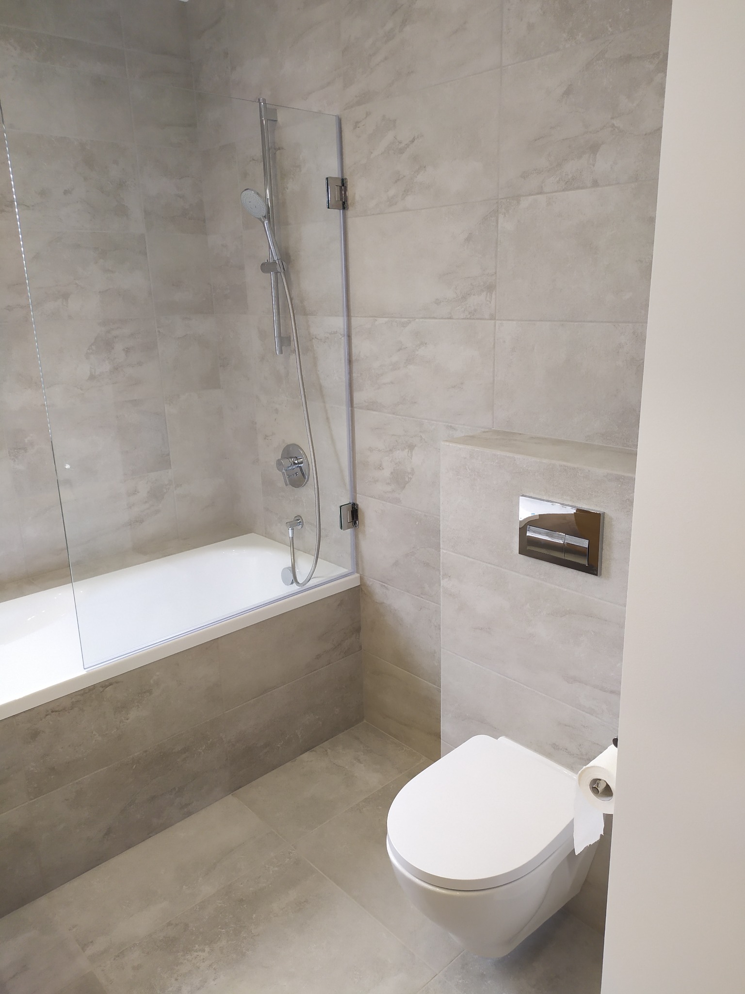 Nowoczesna łazienka w odcieniach szarości z wanną obudowaną płytkami, szklanym parawanem nawannowym, wiszącą toaletą i chromowanym przyciskiem spłuczki.