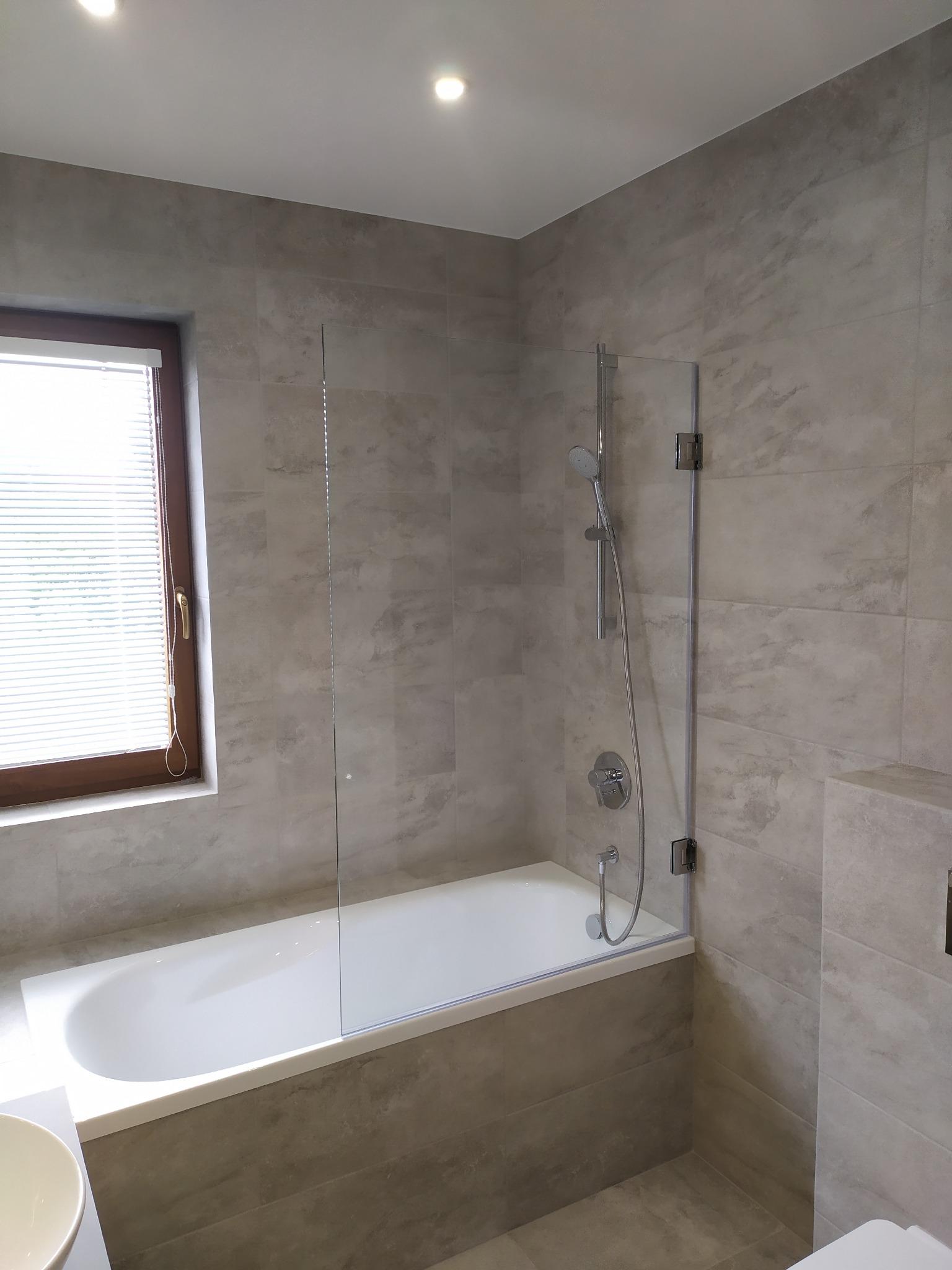 Wykończona łazienka w odcieniach szarości z białą wanną i szklanym parawanem nawannowym z chromowanymi zawiasami, okno z żaluzjami w drewnianej ramie.