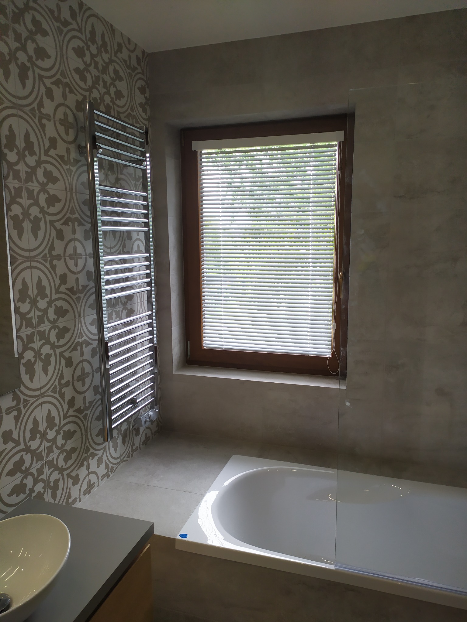 Nowoczesna łazienka z wzorzystymi płytkami, grzejnikiem drabinkowym, oknem z żaluzjami i wanną za szklanym panelem.