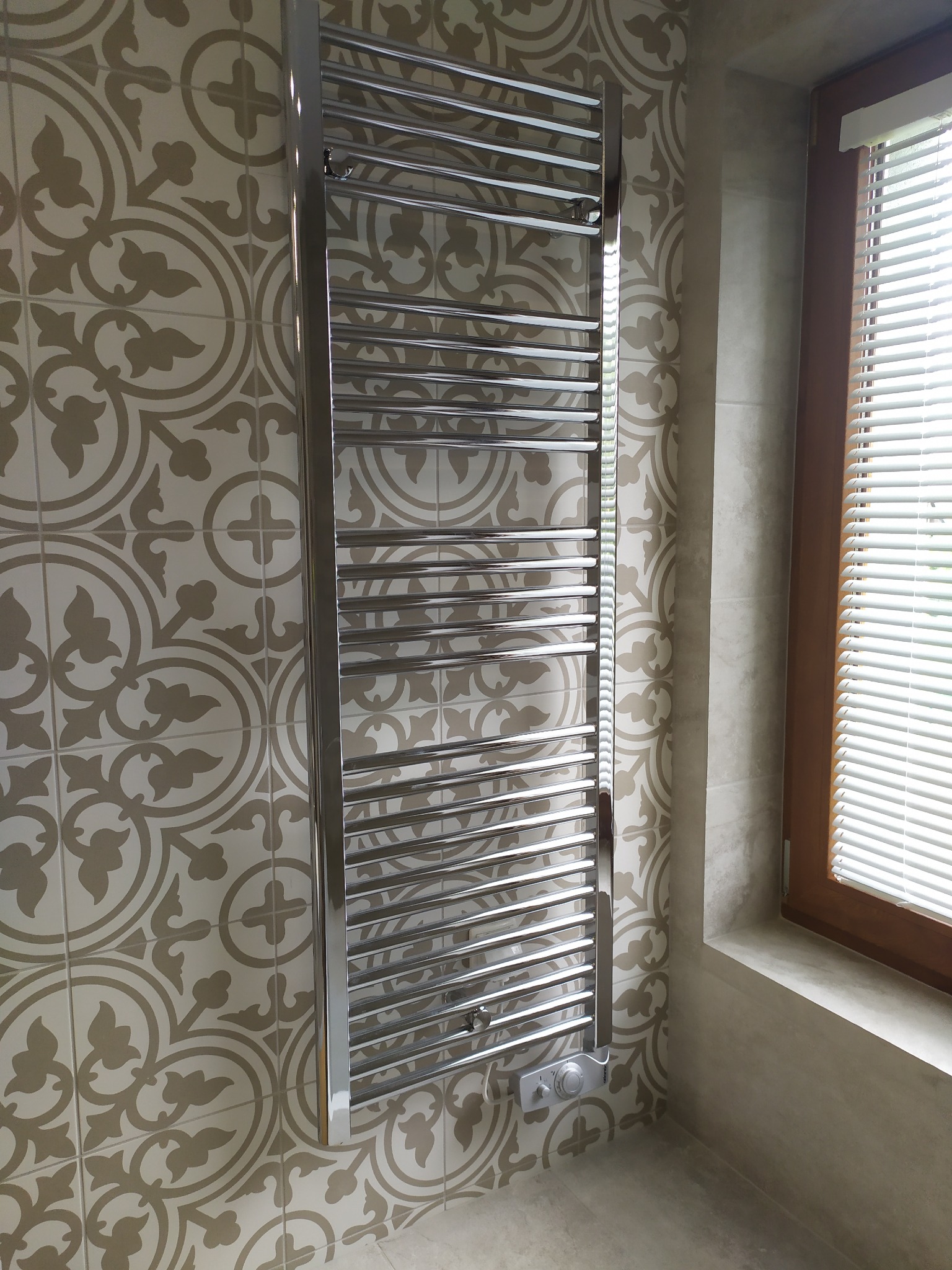 Chromowany grzejnik drabinkowy elektryczny zamontowany na ścianie z dekoracyjnymi, beżowymi płytkami w łazience, obok okna z żaluzjami.