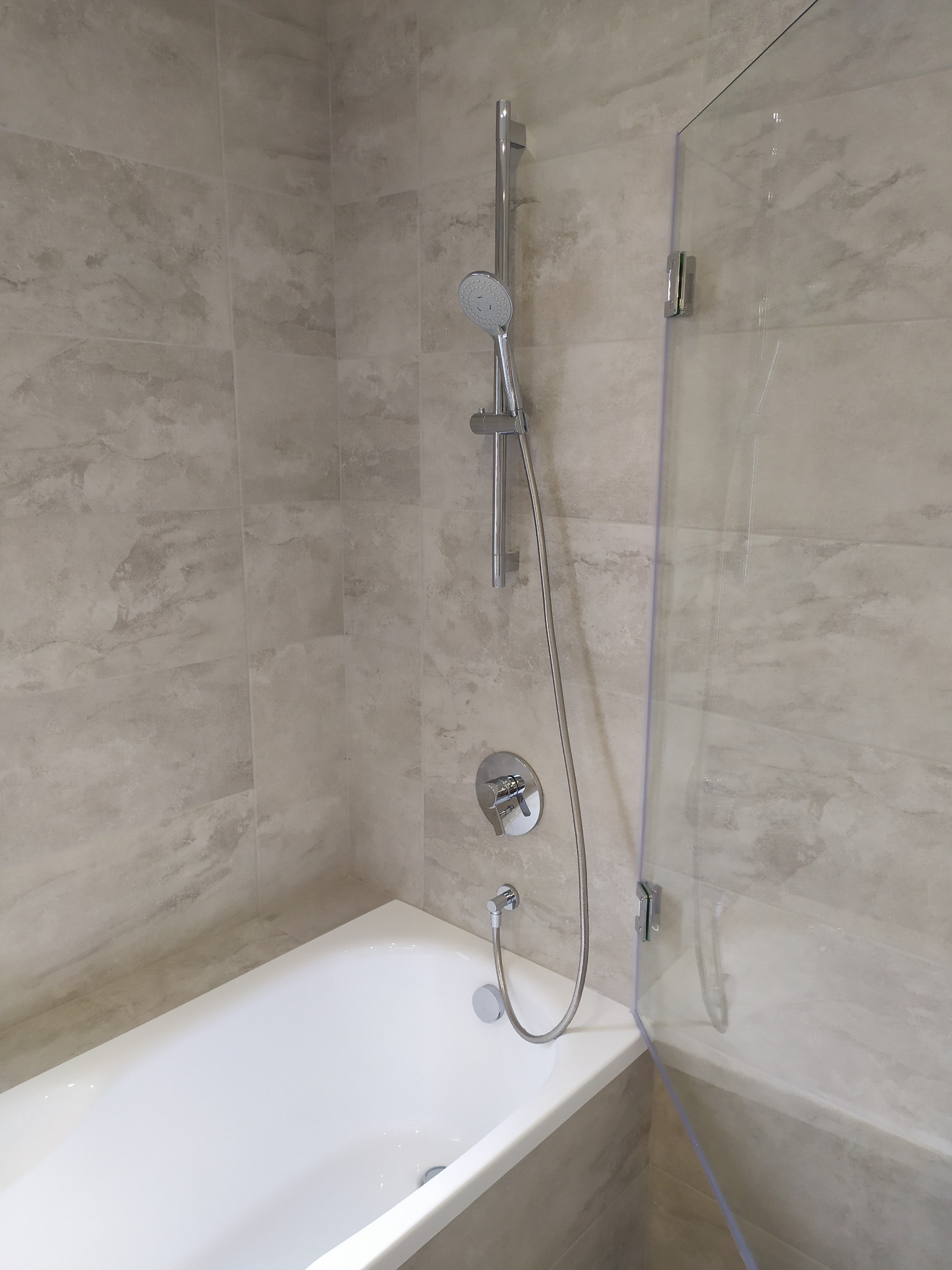 Nowoczesna łazienka z wanną obudowaną beżowymi płytkami imitującymi marmur, szklanym parawanem nawannowym i chromowaną armaturą.